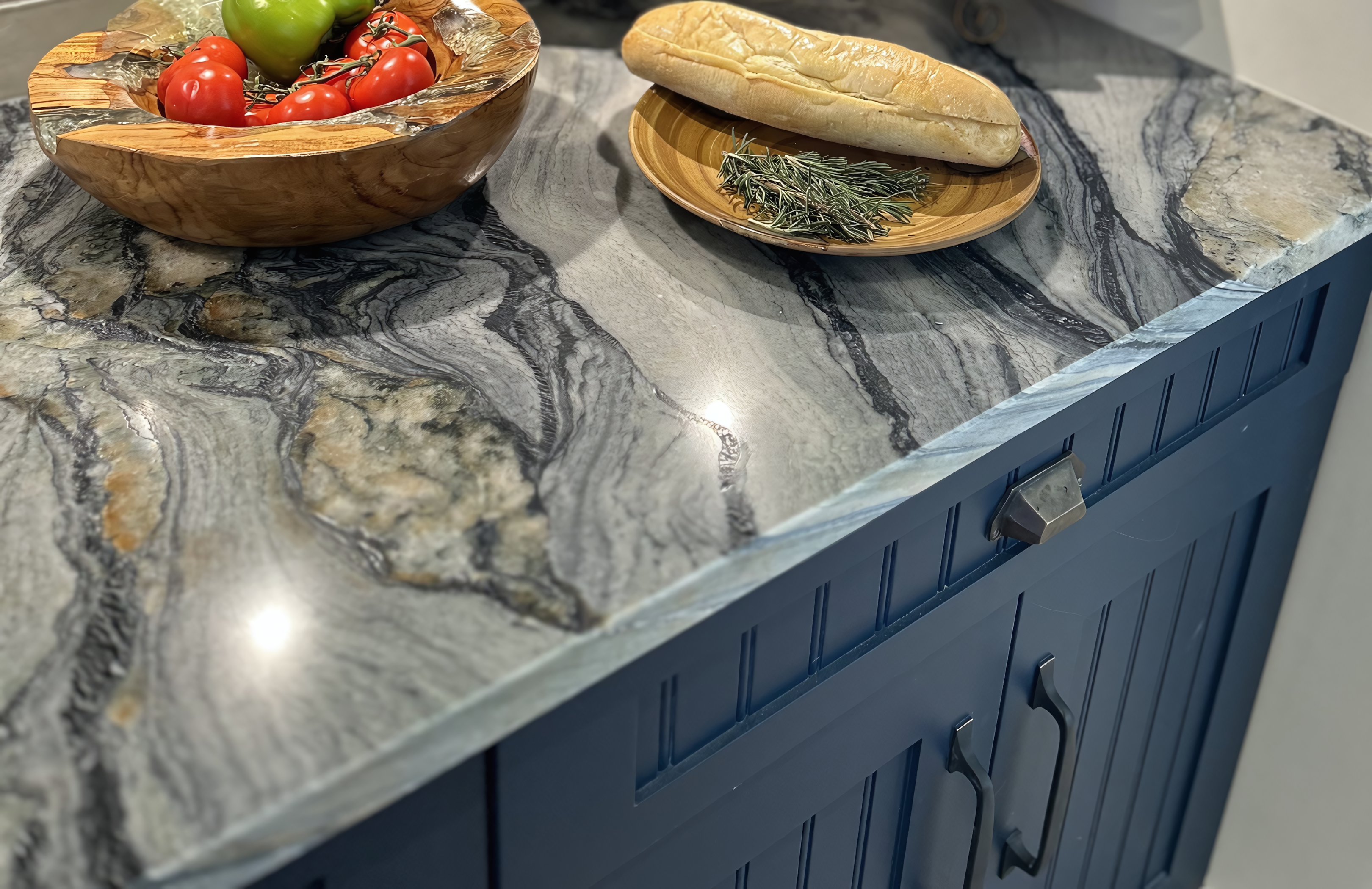 This detailed shot highlights the high-contrast pairing of a natural stone countertop and professional-grade lacquer cabinetry. The countertop features a dramatic, large-scale quartzite slab with deep charcoal and silver veining, creating a sculptural surface for food preparation. Below, the base cabinetry is finished in a deep navy blue lacquer with custom beadboard paneling and integrated micro-shaker details. The look is completed with oil-rubbed bronze cup pulls and a handcrafted live-edge wooden bowl, showcasing a refined intersection of organic materials and precision architectural millwork.