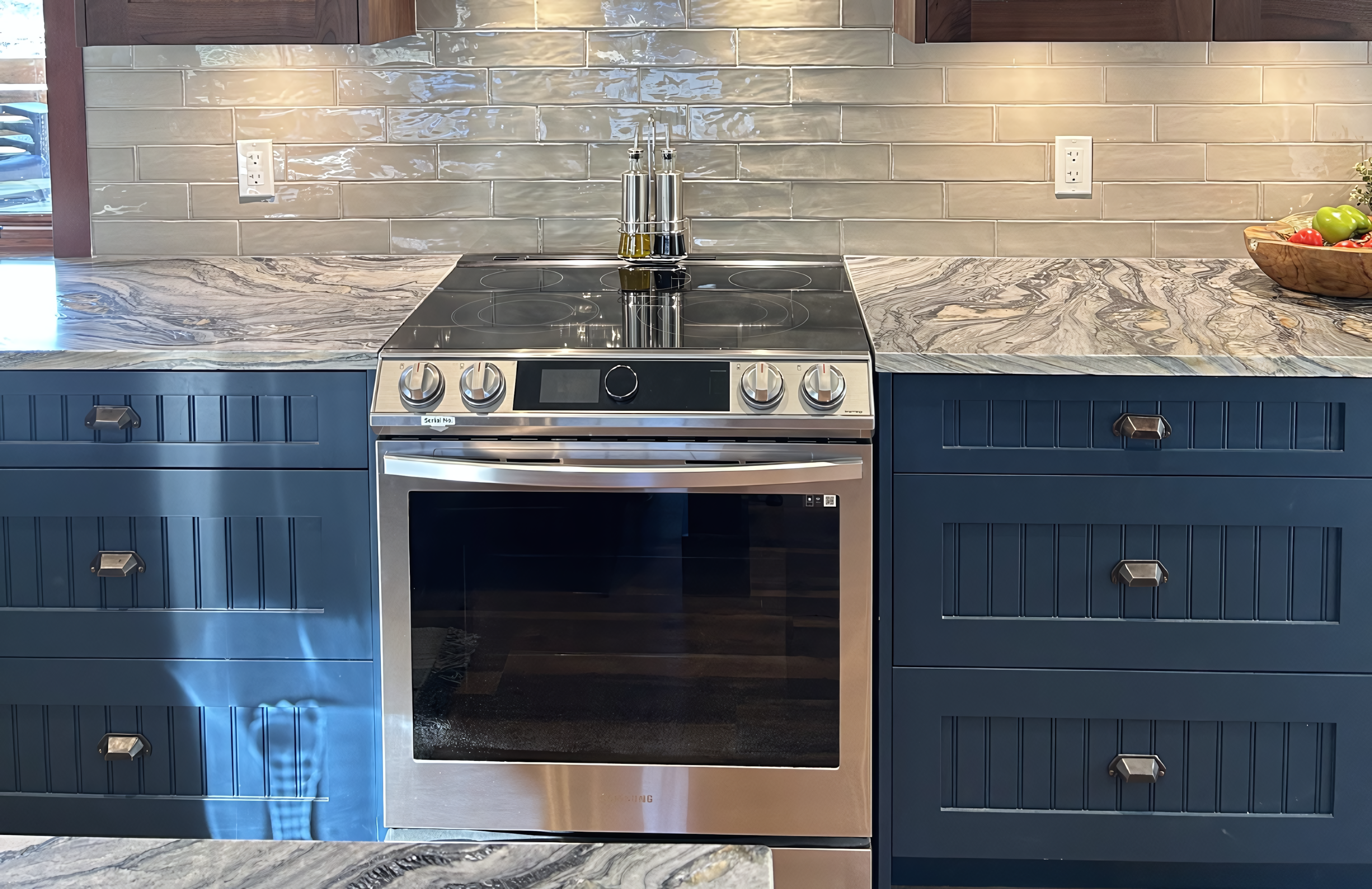 This centered cooking station showcases deep navy blue lacquer base cabinetry with integrated banks of drawers featuring custom beadboard paneling. A slide-in stainless steel range is perfectly fitted between the cabinets, topped by a continuous quartzite countertop with dramatic, flowing grey and gold veining. The design is framed by a glossy, light-grey subway tile backsplash and accented with oil-rubbed bronze cup pulls, demonstrating a precise, professional-grade millwork installation that balances high-performance utility with a refined aesthetic.