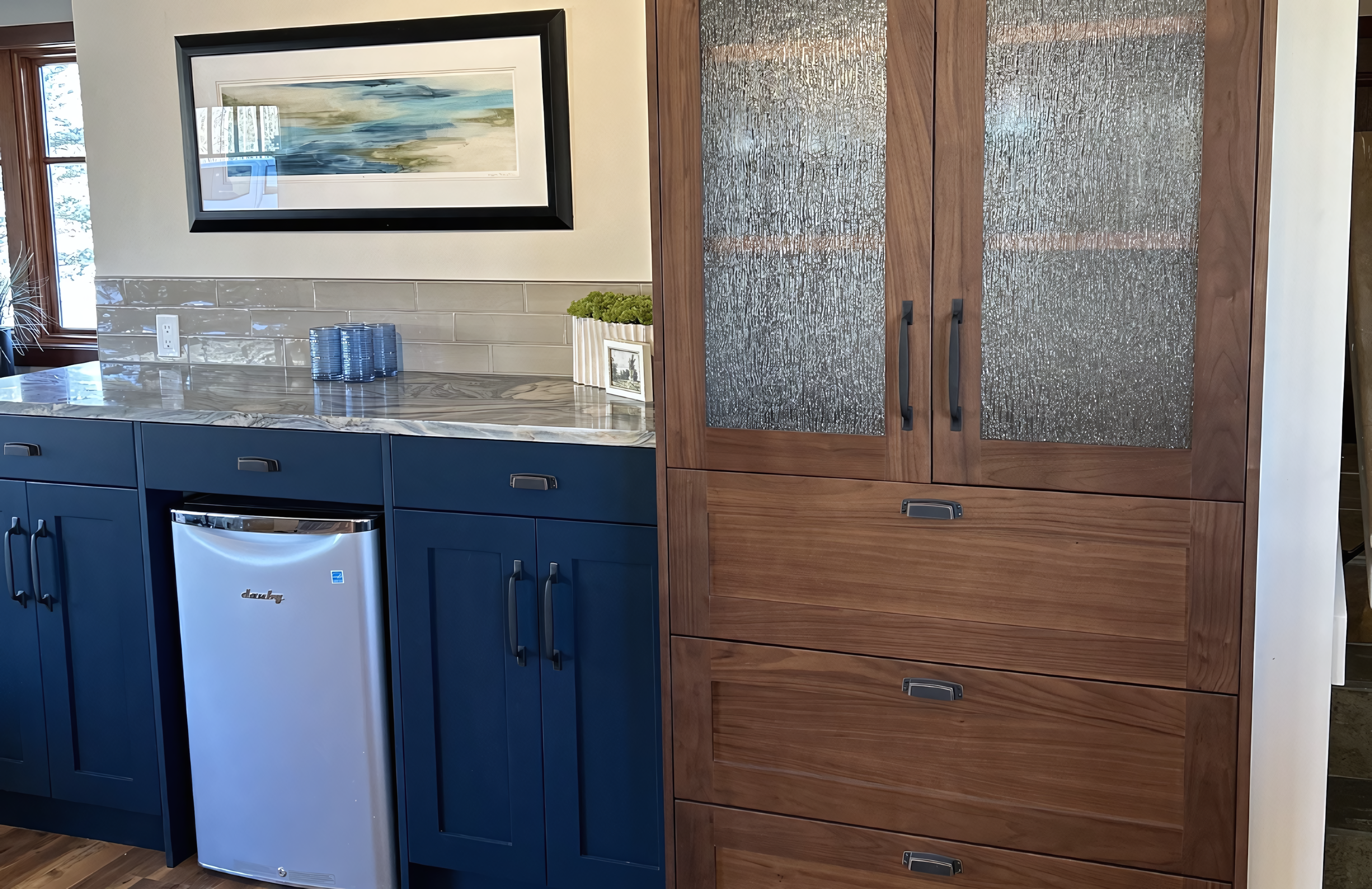 This high-end cabinetry installation continues the tuxedo-style theme, featuring navy blue shaker-style base cabinets that seamlessly integrate a stainless steel beverage refrigerator. Adjacent is a full-height walnut storage unit, designed with a large bank of deep drawers and upper display cabinets featuring textured reeded glass inserts and slim oil-rubbed bronze pulls. The setup is tied together by a consistent quartzite countertop with dramatic veining and a glossy subway tile backsplash, showcasing a refined blend of professional lacquer finishes and natural wood-grain millwork.