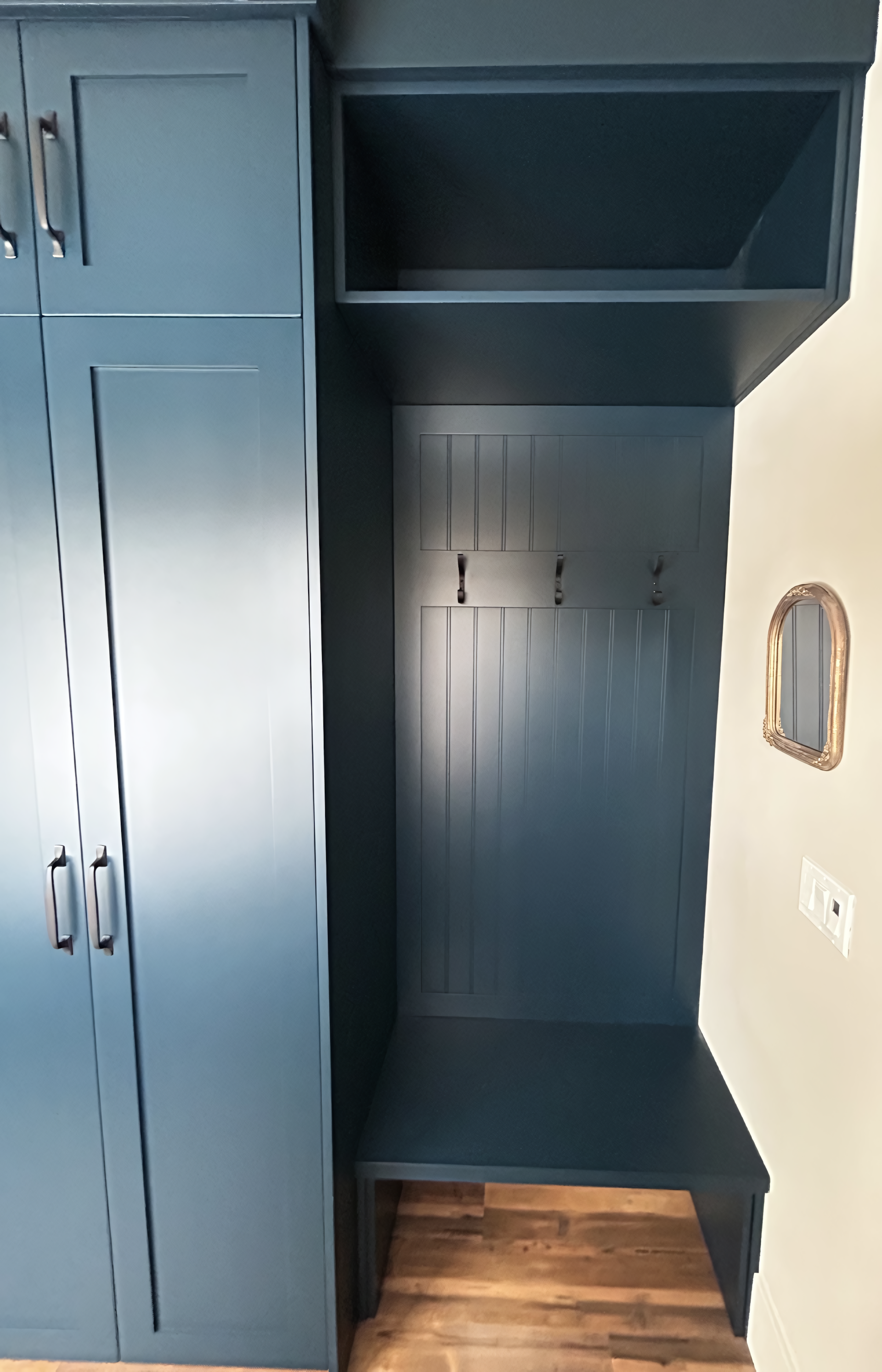Custom built-in entryway storage in a deep blue finish, featuring full-height shaker cabinetry, an integrated bench, vertical back paneling with hooks, and an upper open shelf for organized mudroom functionality.