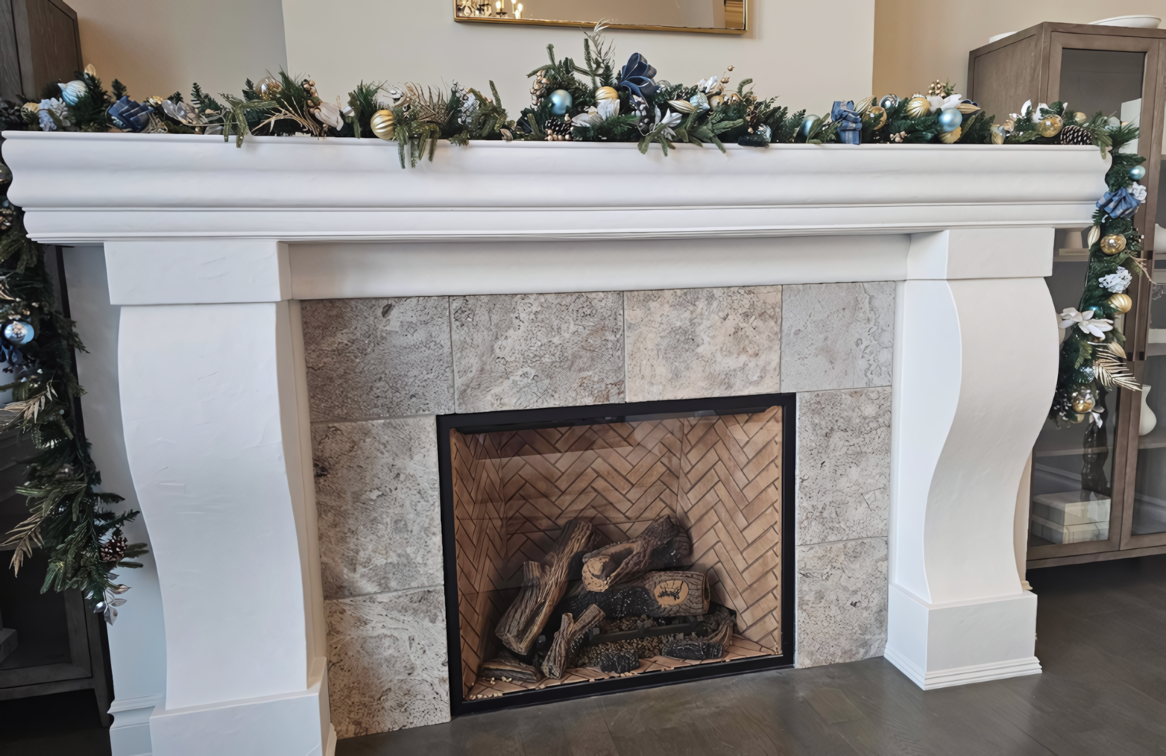 A stately traditional fireplace mantel featuring custom architectural millwork with elegant curved corbel supports and a tiered crown moulding shelf. The white lacquer finish provides a clean, professional aesthetic that contrasts with the natural stone tile surround and herringbone-patterned firebox interior. This bespoke feature piece demonstrates high-end joinery and integrated plinth block transitions at the base for a seamless floor-to-mantel installation.