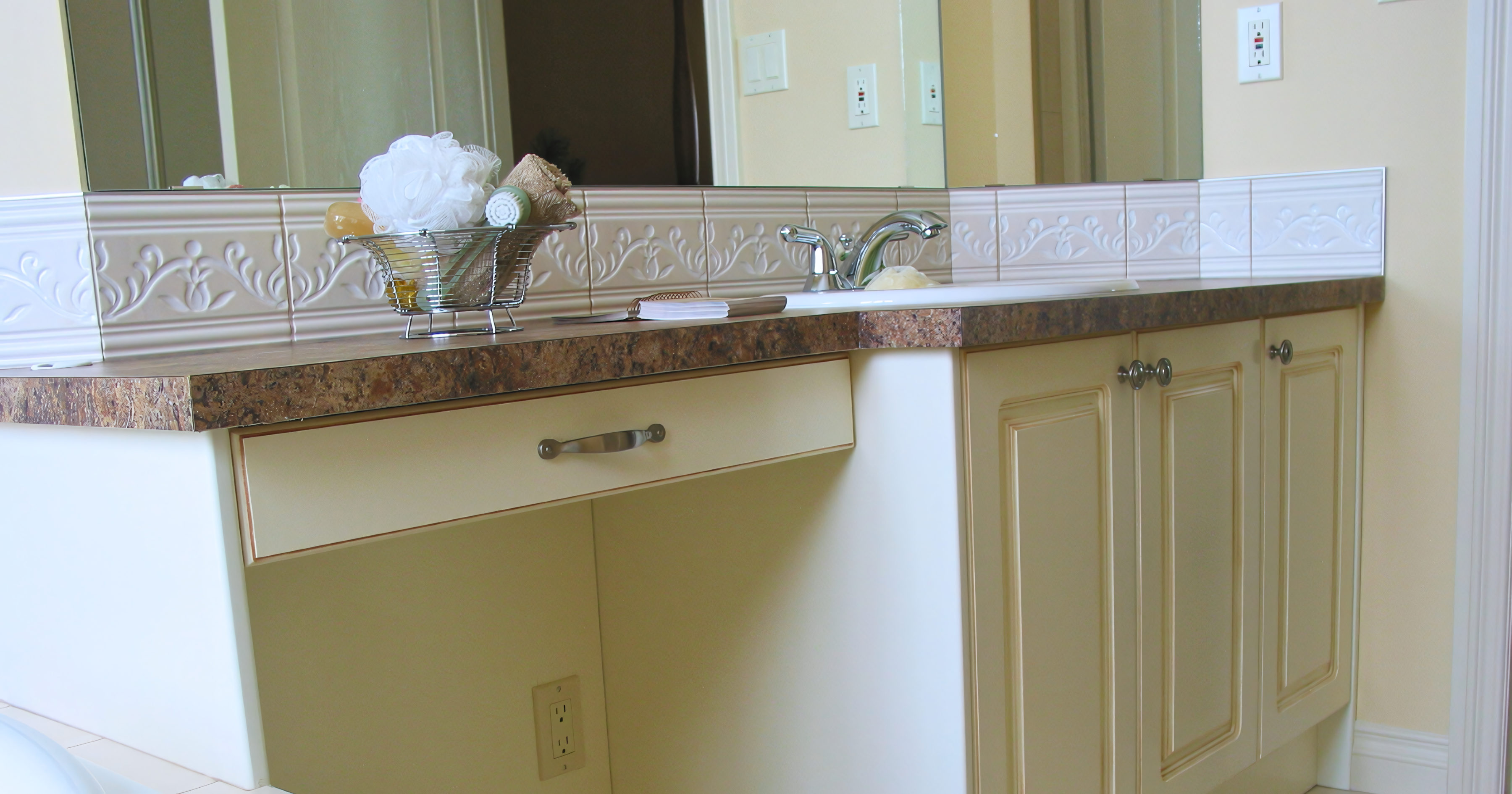 Classic built-in bathroom vanity featuring an off-white or cream painted finish with a subtle brown glaze on the recessed door panels. The design includes a lowered seated grooming station with a single wide drawer, transitioning into a higher section with dual-door cabinetry and a drop-in sink. The unit is topped with a brown granite-look laminate or stone countertop and is accented by a decorative embossed tile backsplash and antique bronze hardware.