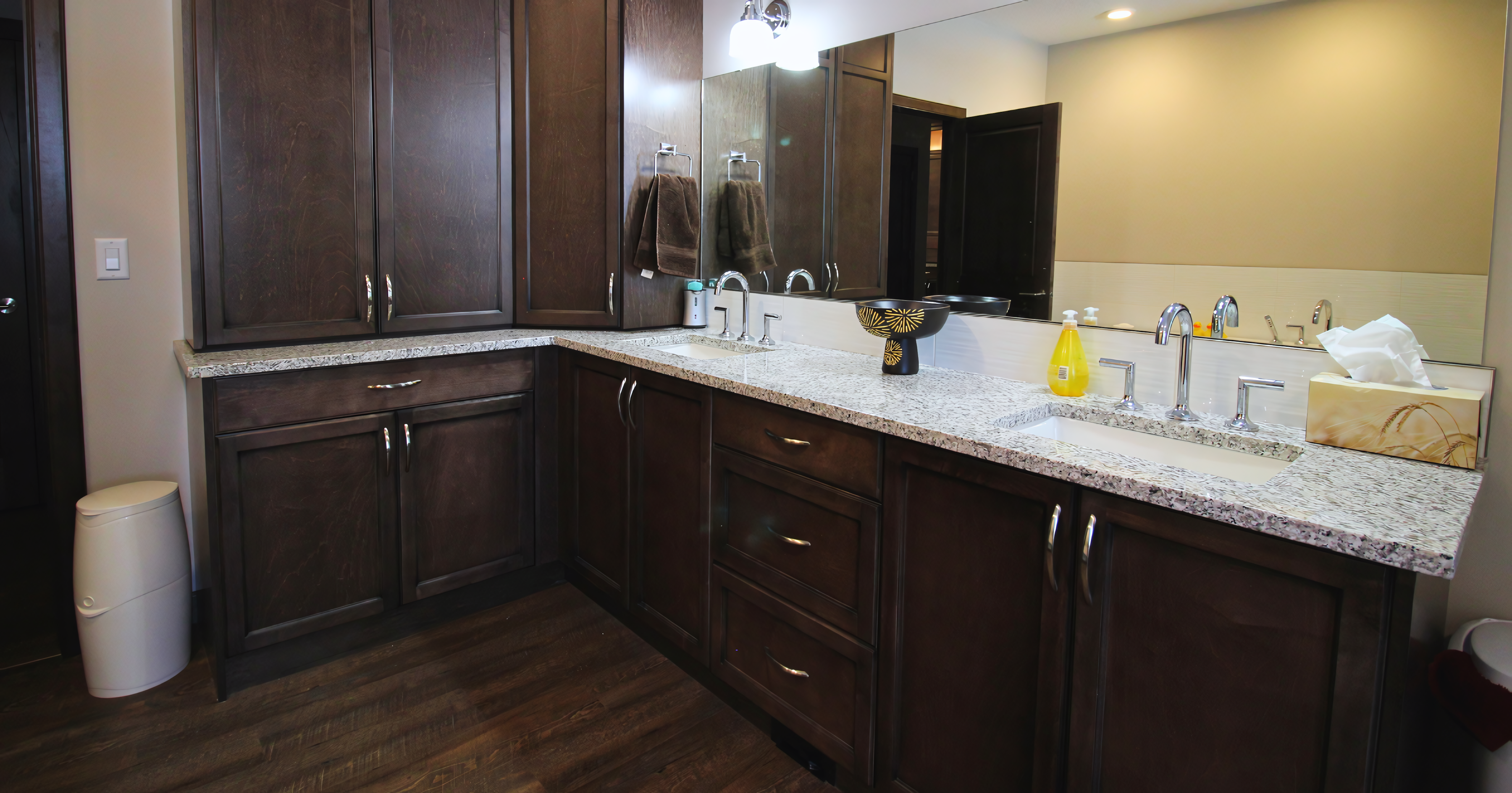 Spacious L-shaped bathroom vanity featuring dark espresso-stained shaker cabinetry with a full-height linen tower for maximum storage. The design includes a speckled granite countertop with dual undermount sinks, polished chrome widespread faucets, and a mix of cabinet doors and drawers equipped with arched satin nickel pulls. The cabinetry is finished with a matching toe kick and sits on dark wood-look flooring, complemented by a large frameless vanity mirror.