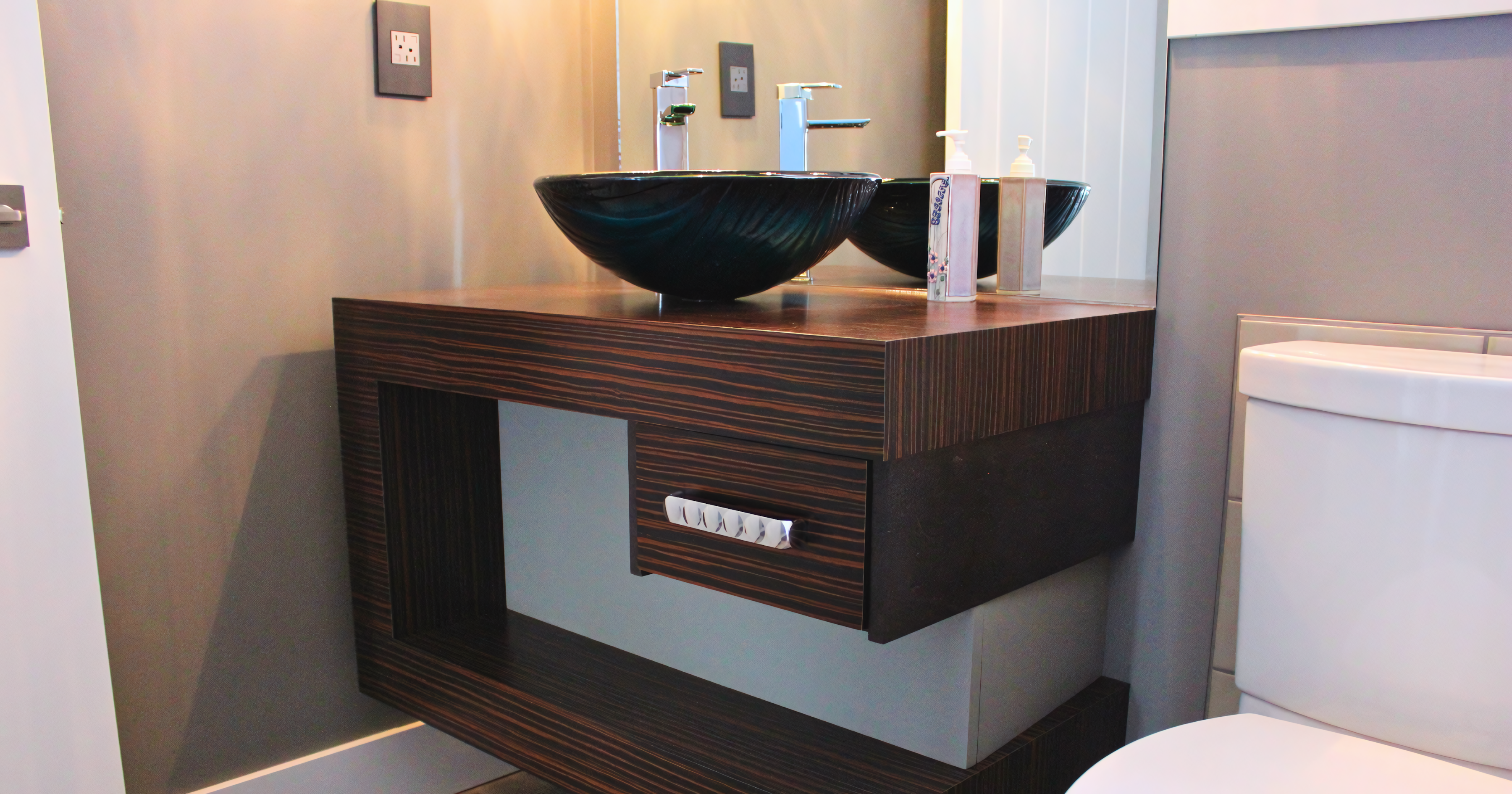 Modern floating bathroom vanity featuring a minimalist asymmetric design with a thick mitered waterfall frame in a dark, linear-grain exotic wood veneer. The unit includes a single offset slab-front drawer with a decorative chrome handle, an integrated open cubby for towel storage, and a black glass vessel sink with a polished chrome faucet set against a full-width mirror.