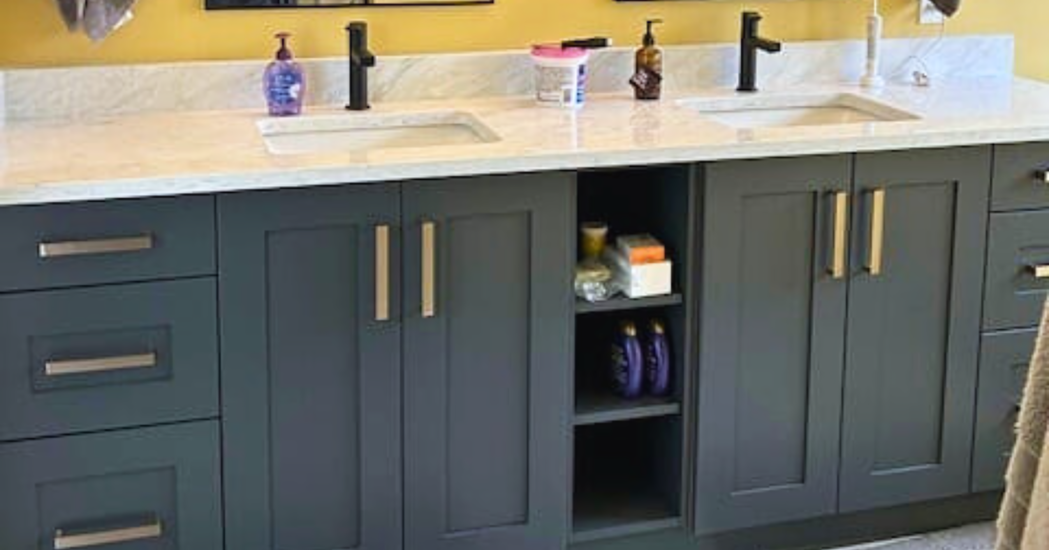 Contemporary double vanity featuring charcoal grey or deep navy shaker-style cabinetry with a professional matte lacquer finish. The layout includes dual undermount sinks set into a white marble-look quartz countertop with a matching backsplash and matte black faucets. The storage configuration consists of two large sink bases, a central open shelving unit for accessible storage, and outer banks of three drawers, all accented with modern brushed gold or brass bar pulls. The cabinetry is finished with a matching recessed toe kick against a mustard yellow wall.