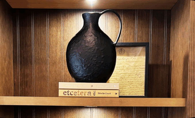 Warm wood open shelving styled with a textured dark charcoal ceramic vase, a stack of books, and a simple framed art piece.