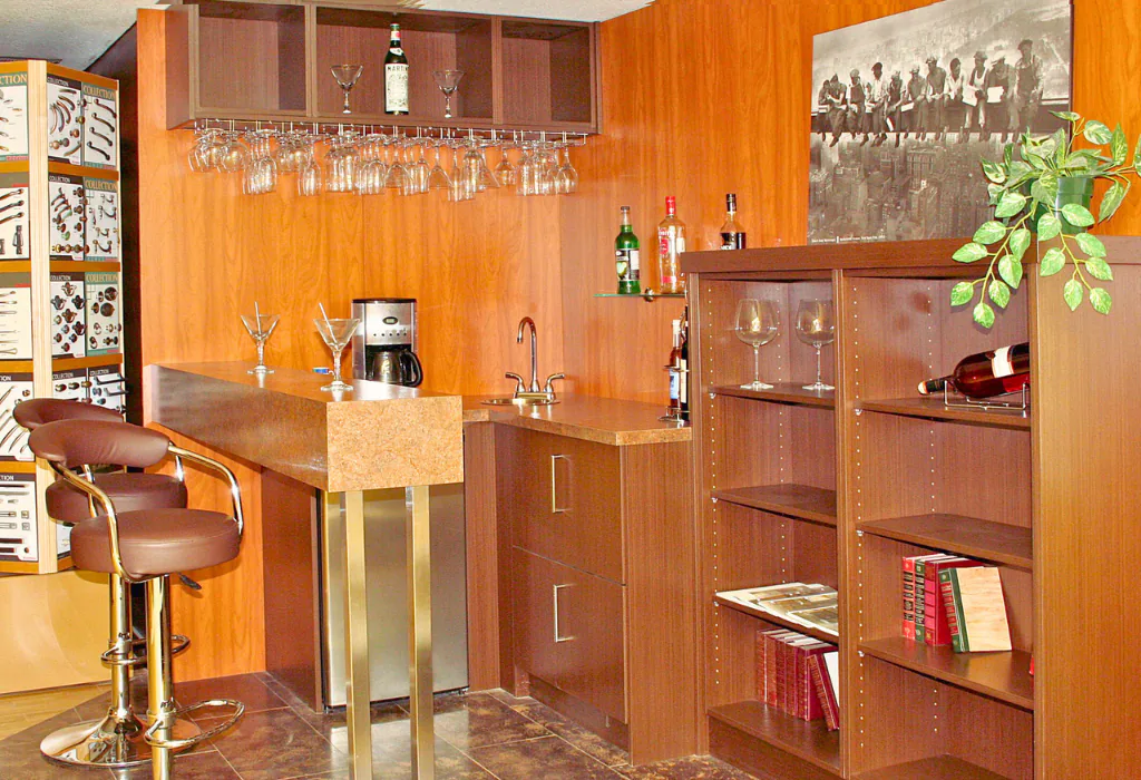 Warm traditional home bar with rich wood-grain cabinetry, integrated stemware rack, full-height matching wood paneling, and a raised bar counter with barstools.