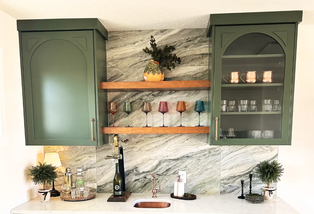 Elegant home wet bar featuring custom sage green lacquer upper cabinets with arched bolection moulding, floating natural wood shelves, and a bold stone backsplash.