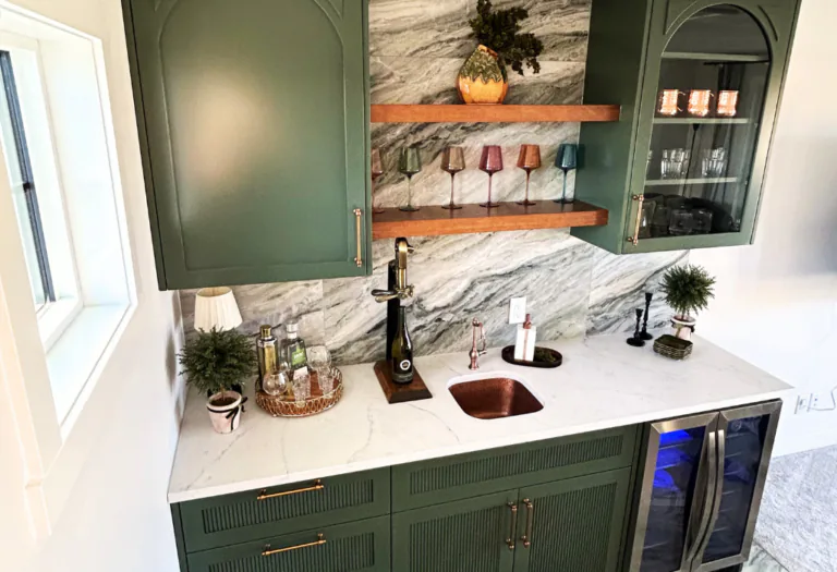 Wide shot of a custom sage green lacquer wet bar featuring arched bolection moulding, floating wood shelves, a fluted tambour base cabinet, and a wrap-around light quartz countertop.