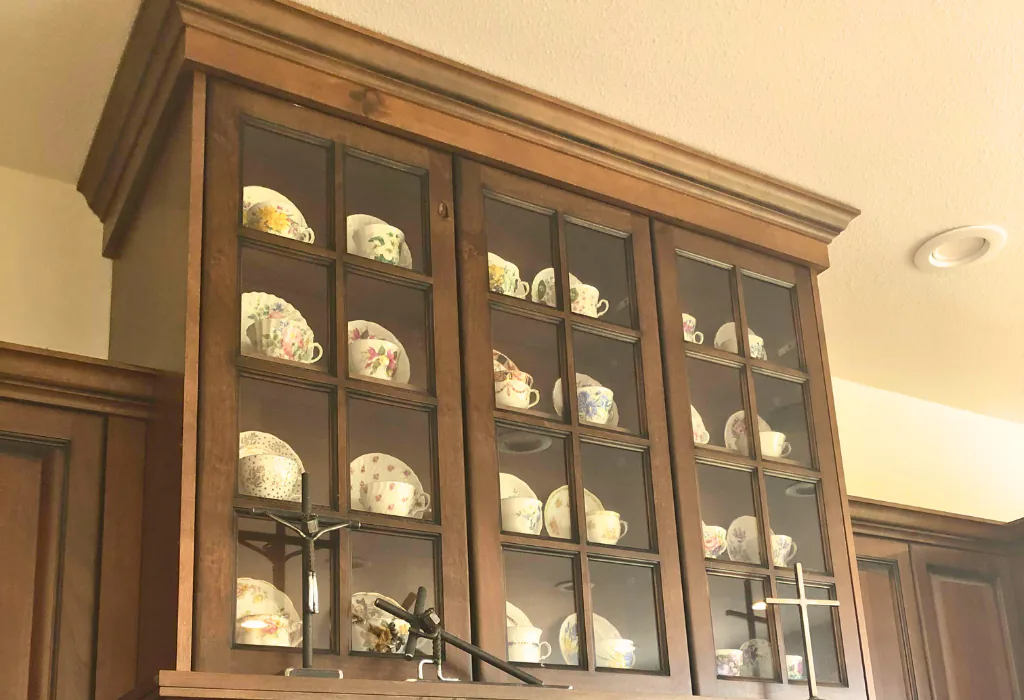 lose-up of an upper-level, warm cherry-stained kitchen hutch featuring three glass-front doors with traditional wood muntin grids showcasing a collection of tea cups.