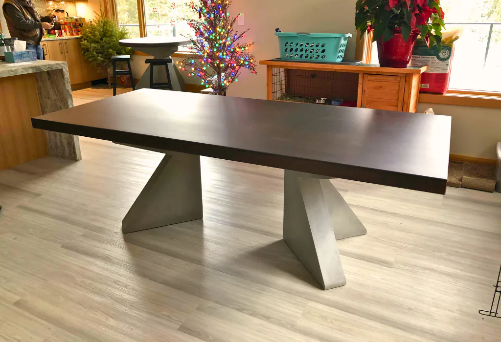 Wide shot of a modern dining table featuring a thick, dark-stained solid wood top supported by two large, geometric, light-grey painted pedestal legs on light wood-plank flooring.