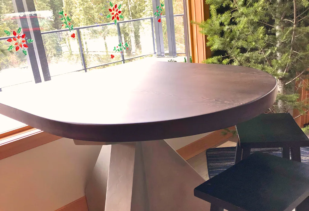 Close-up of a modern round dining table featuring a thick, dark-stained circular wood top and a multi-faceted geometric pedestal base finished in a light grey matte lacquer.