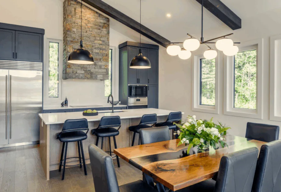 Open-concept kitchen and dining room featuring navy blue shaker cabinets, a large white quartz island with seating, a custom live-edge wood dining table, and vaulted ceilings with dark-stained timber beams.