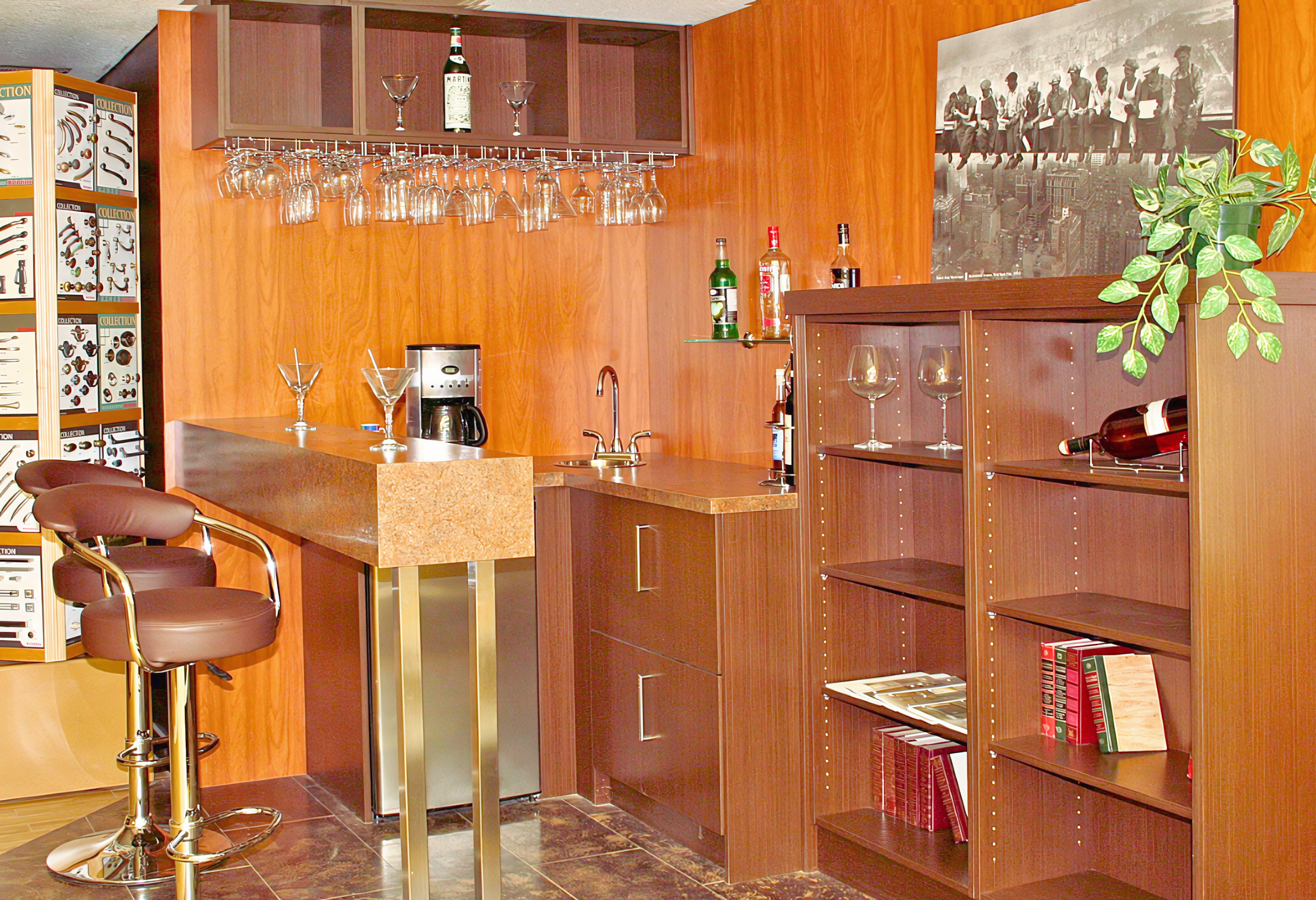 A comprehensive home bar solution featuring flat-panel cabinetry in a warm wood-grain finish with sleek metallic pulls. The design includes a raised peninsula seating area supported by architectural steel legs, alongside a functional prep zone with a small sink and a built-in beverage refrigerator. A matching upper storage unit with an integrated high-capacity stemware rack and adjustable open shelving provides ample display space, while full-height wood wall paneling creates a cohesive, professional millwork aesthetic.
