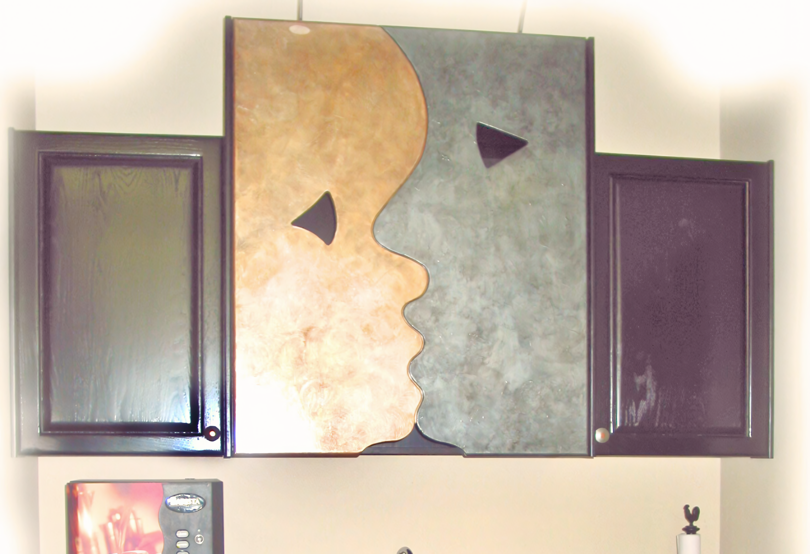 An avant-garde cabinetry installation featuring a custom-carved central focal point with a high-concept "faces" silhouette. The artistic center panels are finished in contrasting metallic liquid-metal coatings—one in warm copper and the other in cool pewter—with integrated triangular cutouts serving as minimalist handles. Flanking the center unit are high-gloss, deep espresso-stained shaker cabinets, demonstrating a unique intersection of fine architectural millwork and specialized decorative finishing techniques.