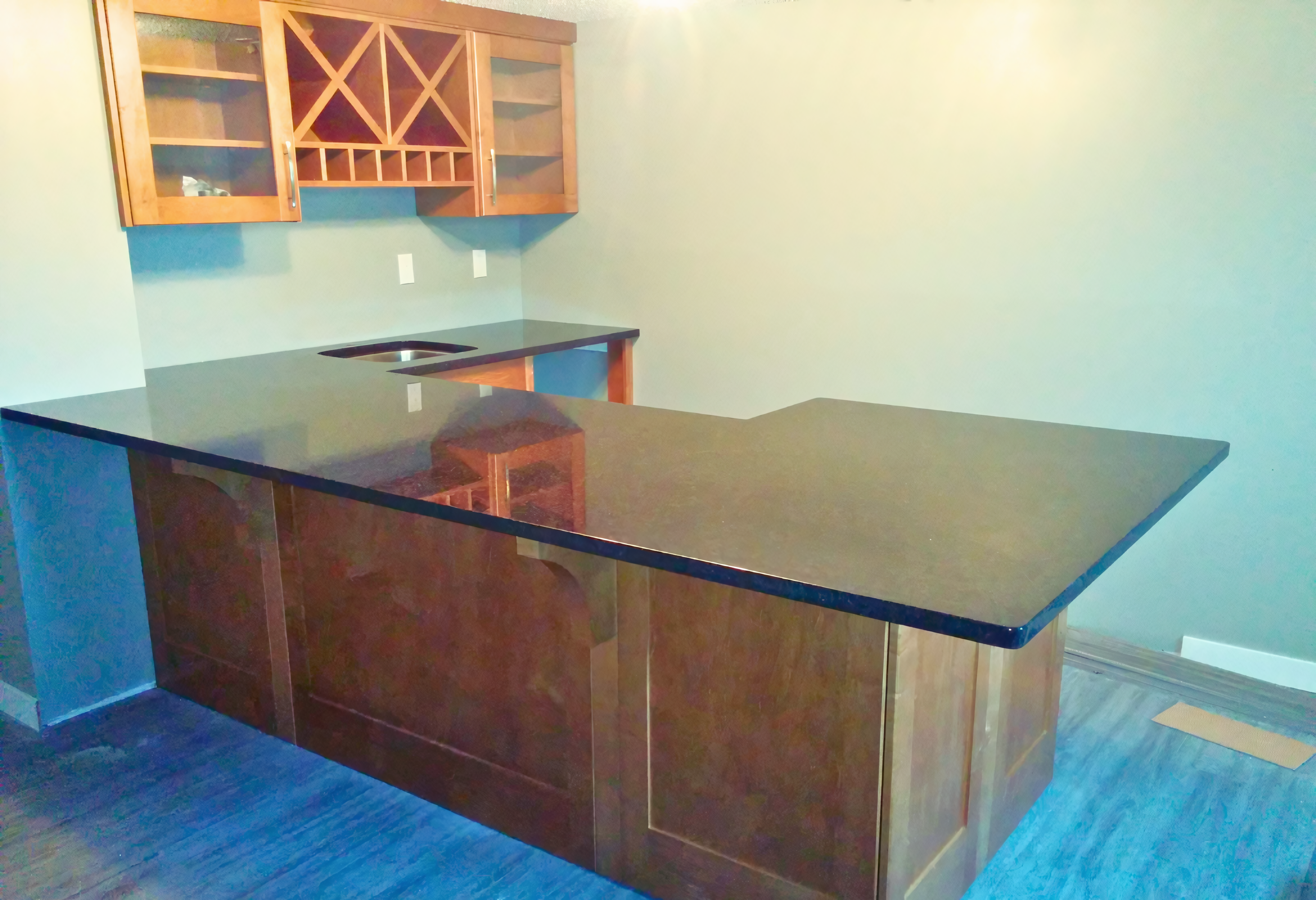 This expansive peninsula-style home bar features warm-toned wood cabinetry with integrated raised panels along the front and a matching upper unit. The upper cabinetry includes a central lattice wine rack and a secondary horizontal bottle storage section, flanked by glass-mullion display doors. A sleek, high-gloss black countertop with a deep overhang provides ample seating area for guests, while the back counter houses an undermount prep sink, showcasing a complete architectural millwork solution for a dedicated entertainment zone.