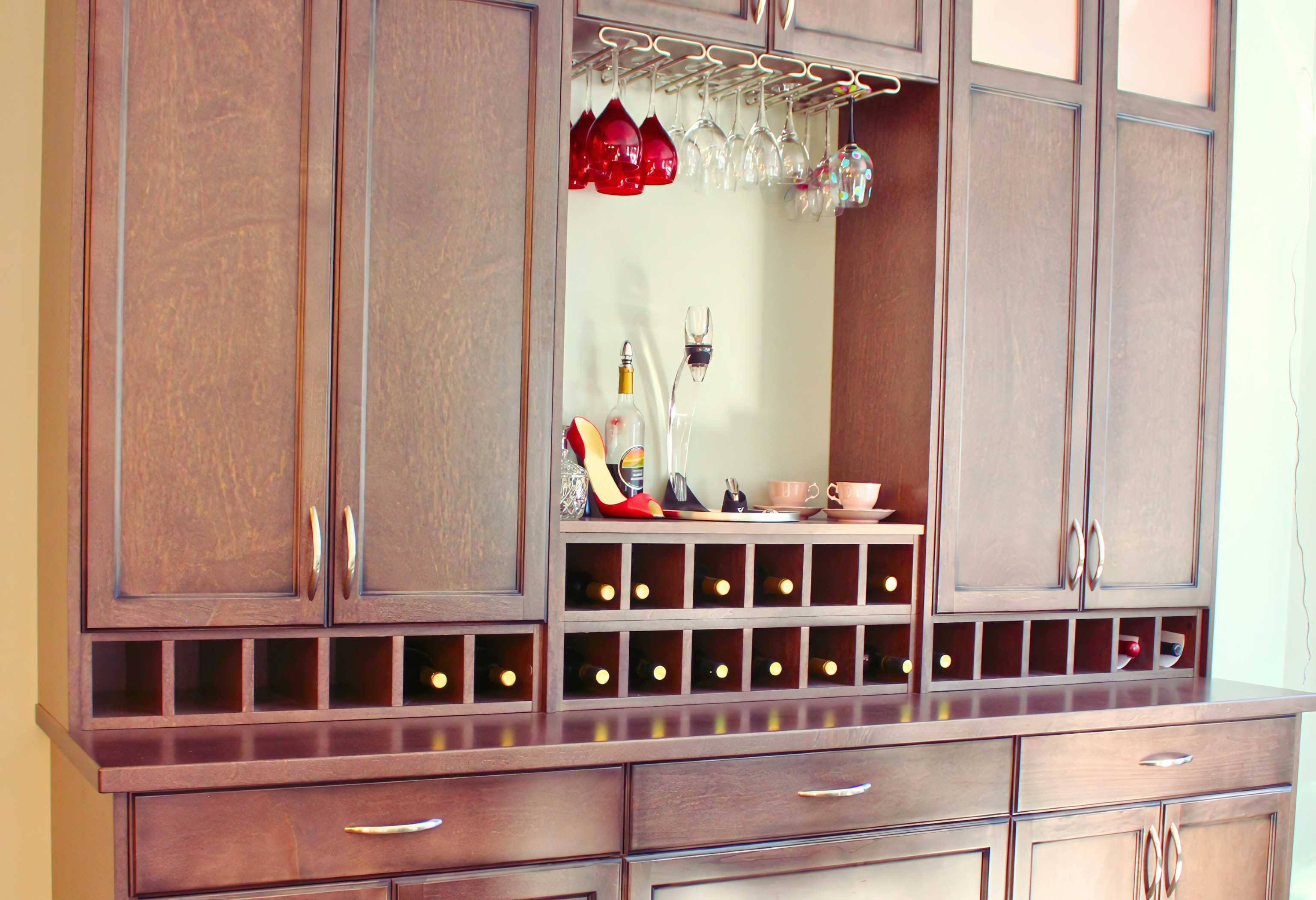 A comprehensive custom home bar and hutch assembly featuring a rich espresso-stained wood finish with shaker-style door panels. The design incorporates an extensive built-in wine cubby system spanning the width of the unit, directly beneath upper storage cabinets and a central bar niche. This functional entertainment hub is equipped with an integrated under-cabinet stemware rack, sleek brushed nickel pulls, and a matching solid-surface countertop, demonstrating high-capacity storage within a refined architectural millwork framework.