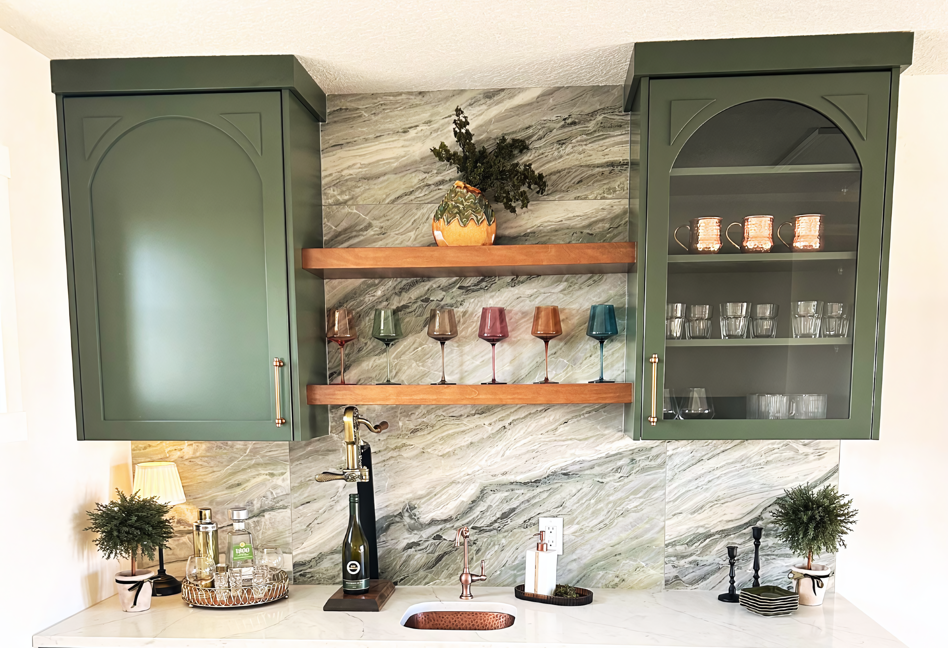 A sophisticated upper cabinetry configuration for a home bar featuring custom sage green lacquer doors with elegant arched bolection moulding details. The layout includes a solid-panel cabinet and a matching glass-front unit for stemware display, flanking a pair of heavy-duty natural wood floating shelves. The backdrop is a dramatic, full-height stone backsplash with sweeping horizontal veining, perfectly complemented by a white quartz countertop and an undermount hammered copper prep sink.