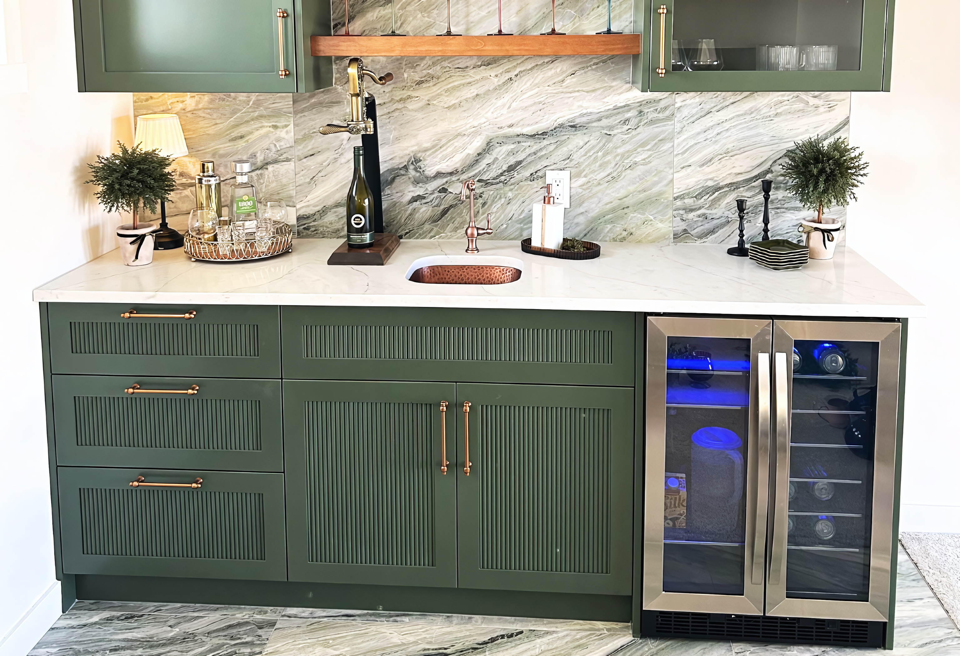 A luxurious custom home bar featuring sage green cabinetry with sophisticated fluted tambour door inserts and gold-toned hardware. The design integrates a professional-grade dual-zone beverage refrigerator alongside a white quartz countertop and an undermount hammered copper sink. A full-height natural stone backsplash with dramatic sweeping veins is accented by a warm wood floating shelf and integrated upper display cabinets, creating a seamless and high-end social entertainment space.